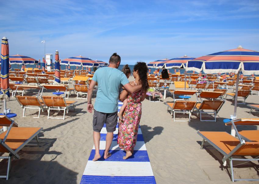 Famiglia cammina sulla spiaggia, sole, ombrelloni colorati e mare all'orizzonte.